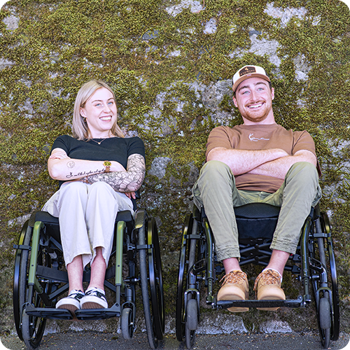 Una giovane donna e un giovanotto sono seduti su carrozzine davanti a un muro di pietra ricoperto di muschio.